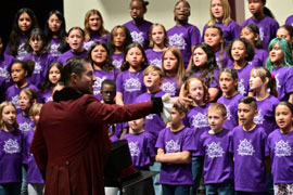 Conductor leading a choir of children wearing purple OMA shirts.