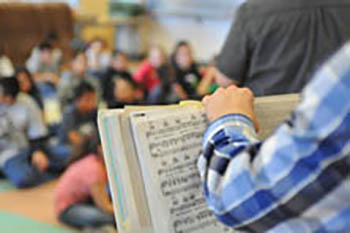 Teacher holds sheet music while a classroom of students listens in the background.