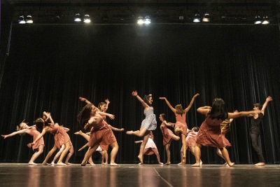 Several dancers perform in flowing dresses on a dark stage under lights.