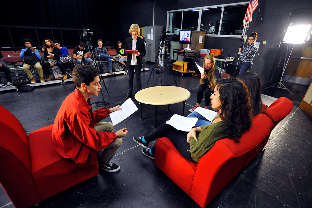 Students rehearse a scene with scripts while sitting in red chairs on a stage.
