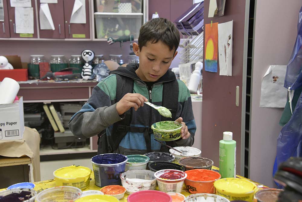 Student mixes paint while standing behind a table covered with containers of different colored paints.